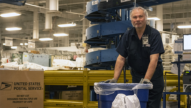 A USPS Laborer Custodial employee holding a recycling receptacle
